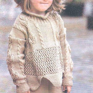 May include: A young girl wearing a knitted beige sweater and skirt with a matching knitted hat. The sweater has a cable knit pattern and a lacy bottom.