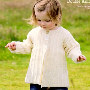 May include: A young child wearing a white knitted cardigan with buttons. The cardigan has a cable knit pattern and is worn over a blue shirt. The child is standing in a grassy field.