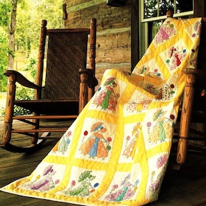 May include: A yellow and white quilt with embroidered figures of women holding parasols. The quilt is draped over a wooden rocking chair on a porch.