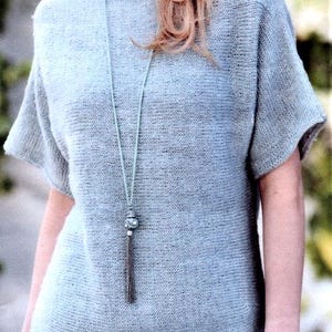 May include: A light blue knit top with short sleeves. The top has a round neckline and a loose fit. A long necklace with a decorative pendant and tassel hangs down the front of the top.