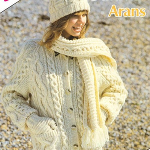 May include: A white knitted cardigan with a cable knit pattern and a matching scarf and hat. The cardigan has a button closure and pockets. The hat has a pompom on top. The text "Outdoor Arans" is visible in the image.