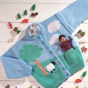 May include: A light blue knitted cardigan with a green, brown, and white landscape design. The cardigan has a pocket on the front with a small knitted shepherd doll inside. The cardigan has a button closure.