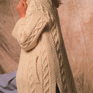 Op de afbeelding: Een crèmekleurige gebreide cardigan met een kabelbreipatroon en een hoge kraag. De cardigan heeft een zijsplit en een zak aan de linkerkant.