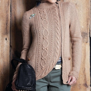 Puede incluir: Un cárdigan de punto trenzado color beige con cuello alto y cierre asimétrico. El suéter presenta intrincados patrones de trenzas y botones de madera. Una mochila negra con tachuelas doradas se sostiene en la mano izquierda. El fondo es una pared de madera.