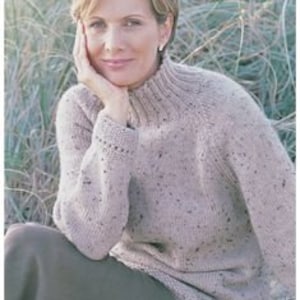 May include: A woman wearing a light brown speckled knit sweater with a high collar and long sleeves. The sweater is labeled "Aran / Worsted Yarn" and sized 32-42 inches. The woman is sitting outdoors with tall grass in the background.