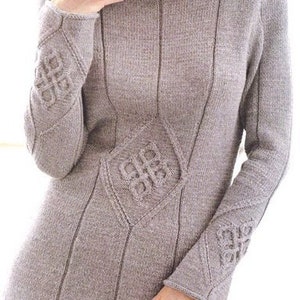 Puede incluir: Un suéter de punto gris con un diseño único que presenta intrincados patrones de nudos en las mangas y el frente. El suéter tiene un ajuste holgado y un escote ligeramente redondeado.