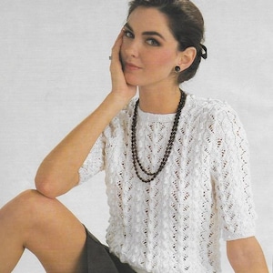 May include: A white crocheted top with a delicate openwork pattern and a round neckline. The top has short sleeves and is worn with a black skirt and a black beaded necklace.