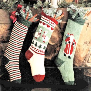 May include: Three knitted Christmas stockings hang in front of a fireplace. One stocking has red and green stripes, another has geese and hearts, and the third features Santa Claus. Each stocking is decorated with green trim and cinnamon sticks.