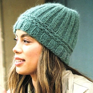 May include: A woman wearing a teal blue knitted beanie hat with a cable knit pattern. The hat is made of a soft, fuzzy yarn.