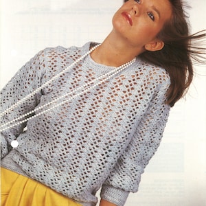 May include: A light blue crochet sweater with a loose fit and a delicate openwork pattern. The sweater is worn over a yellow skirt and the model is wearing a pearl necklace.
