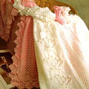 Peut inclure: Deux couvertures pour bébé en crochet, une blanche avec un motif floral et une rose avec un motif en dentelle. Les couvertures sont pliées et drapées sur un panier en osier.