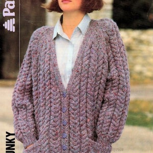 May include: A purple and blue chunky knit cardigan with a cable knit pattern and pockets. The cardigan has a V-neck and buttons down the front. The model is wearing a white button-down shirt underneath.
