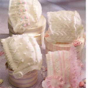 May include: Four white knitted baby bonnets with different patterns and lace trim. The bonnets are displayed on pink and white boxes.