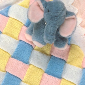 Puede incluir: Una manta de bebé tejida con un patrón tejido en tonos de rosa, azul, amarillo y blanco. Un juguete de peluche gris en forma de elefante con orejas rosas se coloca encima de la manta.