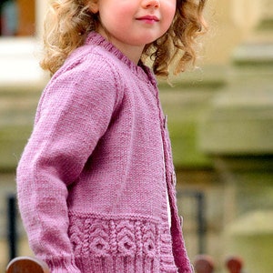 May include: A young girl wearing a pink knitted cardigan with a cable knit pattern on the bottom edge. The cardigan has a button closure and is made of a soft, textured yarn.