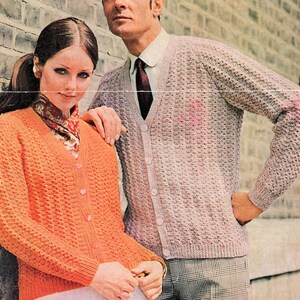 May include: A couple wearing matching knit cardigans. The woman's cardigan is orange and the man's cardigan is light pink. Both cardigans have a V-neck and button closure.
