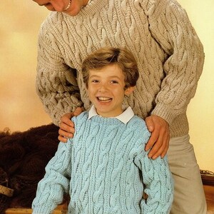 May include: A father and son wearing matching cable knit sweaters. The father is wearing a beige sweater and the son is wearing a light blue sweater.
