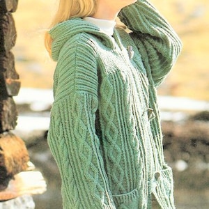 Puede incluir: Un cárdigan verde con capucha, de punto de cable y con bolsillos. El cárdigan está abierto y la capucha está levantada.