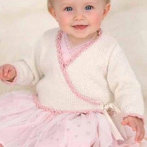 May include: A baby in a white knitted wrap cardigan with pink edging and a pink tulle skirt. The cardigan has a wrap-around style with a tie. The skirt is layered and fluffy, creating a soft, delicate appearance.