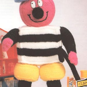 May include: A whimsical, handmade knitted doll with a pink head, black nose, and a smiling mouth. It wears a blue hat, a black and white striped shirt, yellow shorts, and blue shoes. The doll has black arms and a black cane.