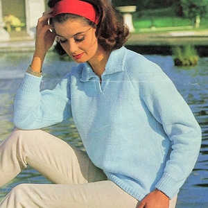 Pode incluir: Um suéter de malha azul claro com gola e fecho de zíper. O suéter tem mangas compridas e um caimento solto. A modelo está usando o suéter com uma faixa de cabeça vermelha e calças bege.