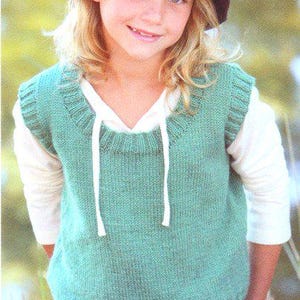 Può includere: Una persona giovane indossa un gilet lavorato a maglia verde acqua con coulisse bianca e un cappello da pescatore grigio scuro. Il gilet è sovrapposto a una camicia bianca a maniche lunghe. La persona sorride.