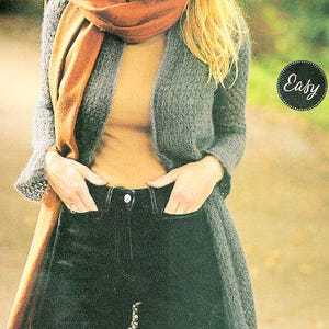 May include: A gray knitted cardigan with a long, open front and a brown scarf. The cardigan is worn over a yellow turtleneck shirt and black jeans.