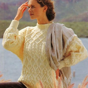 May include: A white knitted sweater with a cable knit pattern. The sweater has a high neck and long sleeves. The model is wearing the sweater with a light brown scarf.