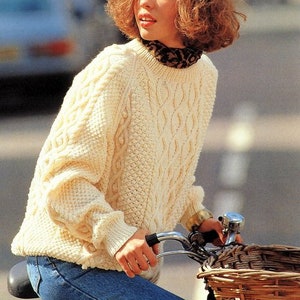 May include: A woman wearing a white cable knit sweater with a brown and black leopard print scarf, riding a bicycle with a wicker basket.