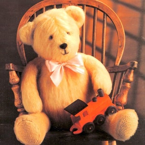 May include: A plush, golden-yellow teddy bear with a pink bow tie sits in a wooden chair. The bear holds a small, orange wooden toy train. The chair has a spindle back.