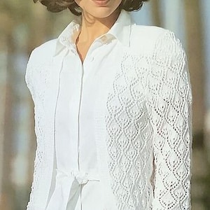 May include: A white crocheted cardigan with a delicate openwork pattern. The cardigan is worn over a white button-down shirt with a tied sash.