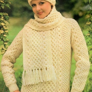 Könnte beinhalten: Ein weißer Strickpullover mit passendem Schal und Mütze. Der Pullover hat ein Zopfmuster und der Schal hat Fransen.