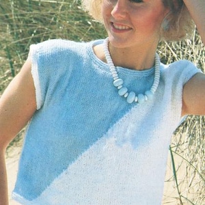 May include: A light blue and white knitted top with a round neckline. The top is sleeveless and has a diagonal color block design. A white beaded necklace with blue accents is worn. The model is smiling.