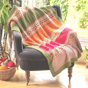 May include: A vibrant, knitted blanket in shades of green, orange, and pink, draped over a dark grey armchair. The blanket displays detailed patterns. A basket of yarn balls is visible, indicating a handcrafted item.