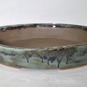 May include: An oval ceramic bonsai pot with a smooth, matte brown interior and a reactive glaze exterior featuring shades of sage green, slate blue, and dark charcoal. The glaze exhibits a distinct drip pattern cascading down the sides.