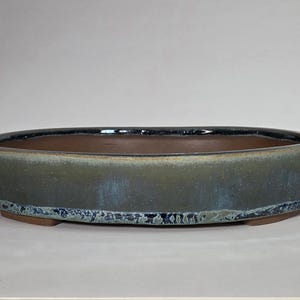 May include: An oval-shaped bonsai pot with a dark brown interior and a glazed exterior. The exterior has a gradient of green and blue hues, with a textured, light blue rim. The pot sits on small, rectangular feet.