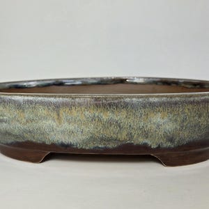 May include: An oval-shaped bonsai pot with a glazed finish. The pot features a dark brown base and a gradient of colors, including green, brown, and blue. The pot is elevated by three small feet and is suitable for bonsai trees.