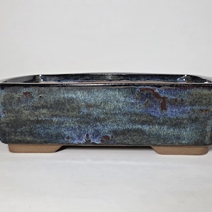 May include: Rectangular bonsai pot with a glazed finish. The ceramic pot has a dark blue and green mottled appearance with a glossy sheen. The base is a light brown color, and the pot is elevated on small feet.