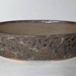 May include: An oval-shaped ceramic bonsai pot with a low profile and a wide opening. The exterior features a textured, mottled glaze in shades of dark brown, tan, and muted green, while the interior has a smooth, light beige finish.
