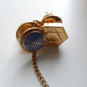 May include: A vintage gold-tone tie tack set. The set includes a round blue and gold emblem, a geometric shape, and a cylindrical piece. A gold chain connects the pieces. The tie tack is on a white surface.