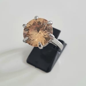 May include: A silver-toned ring featuring a large, round, faceted champagne-coloured gemstone held in place by a six-prong setting. The band is smooth and metallic, showcasing a classic solitaire design with a prominent, light-brown centre stone.
