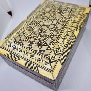Pode incluir: Uma caixa retangular de madeira com um intrincado design embutido. A tampa apresenta um padrão geométrico de triângulos, quadrados e estrelas em tons de creme, preto e castanho. A caixa tem uma borda creme e uma base castanho escuro.