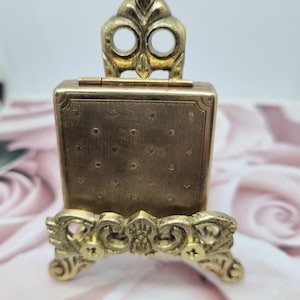 Puede incluir: Un estuche compacto rectangular de metal dorado con una superficie texturizada que presenta un patrón de pequeños puntos y estrellas. El compacto se exhibe sobre un soporte decorativo de metal dorado con volutas ornamentadas y dos tornillos visibles.