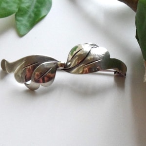 May include: A silver-coloured leaf brooch with a stylised design. The brooch features three sets of leaves, with a pin on the back for fastening. The leaves have a smooth, reflective surface, and the brooch is likely made of metal.