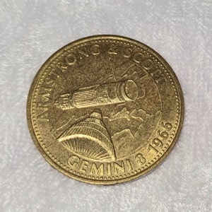 May include: A gold-colored coin with the text "Armstrong & Scott Gemini 8 1966" around the edge. The center of the coin features a depiction of a spacecraft.