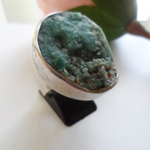 May include: A silver ring featuring a large, green, irregularly shaped gemstone. The gemstone has a rough, textured surface.