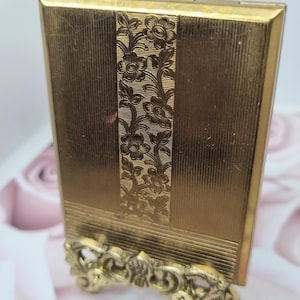 Op de afbeelding: Een rechthoekig goudkleurig metalen compactdoosje met een verticaal middenpaneel met een reliëf bloemmotief en fijne horizontale lijntexturen op de zijpanelen, rustend op een decoratieve goudkleurige metalen displaystandaard.