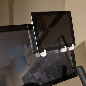 May include: A white tablet holder attached to a black monitor. The tablet holder has three white hooks that hold the tablet in place.