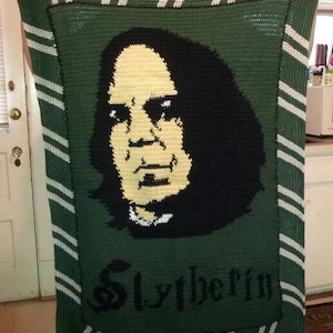 May include: A green crocheted blanket with a white and green striped border. The blanket features a portrait of a man with black hair and a yellow face. The text "Slytherin" is written in black letters at the bottom of the blanket.