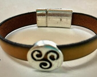 May include: A brown leather bracelet with a silver magnetic clasp and a silver charm featuring a stylized triple spiral design.
